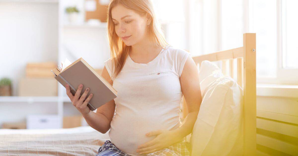 a pregnant woman wearing a white top, reading a book and cradling her bump