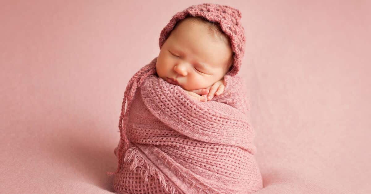 A newborn baby sleeps peacefully while swaddled in a soft pink knitted wrap, wearing a matching pink bonnet. The baby is propped upright against a pink background, with their hands tucked gently under their chin.