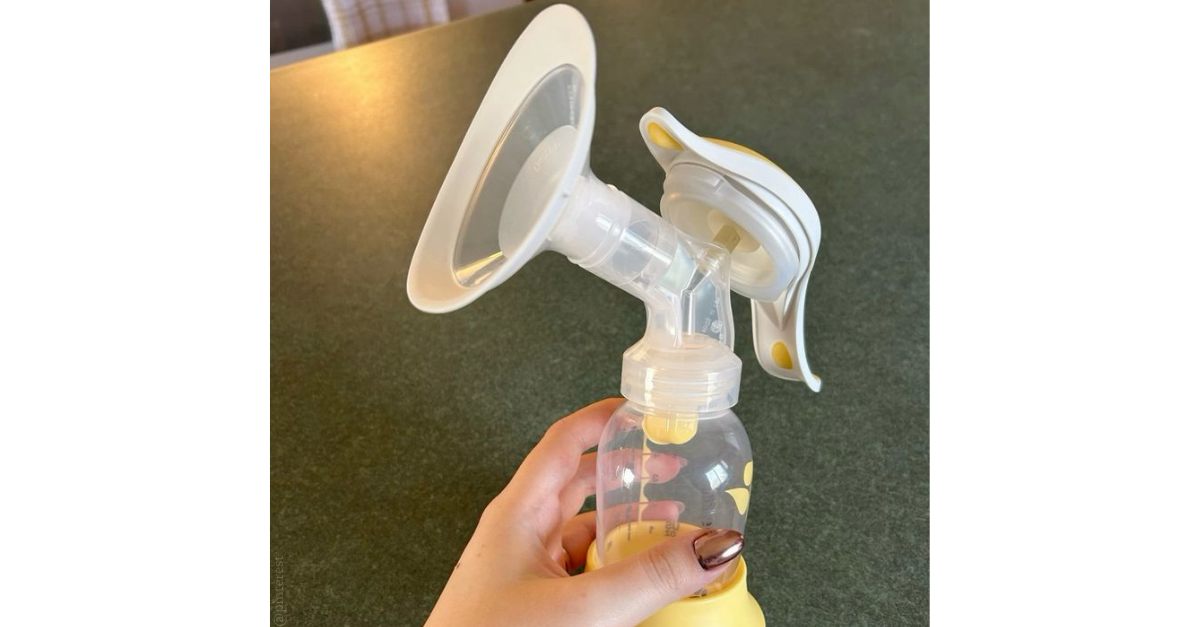 Hand holding a manual breast pump with clear breast shield and yellow-handled lever over a kitchen-style worktop.