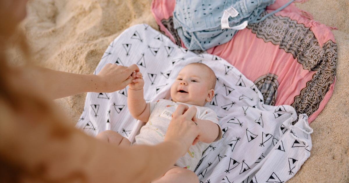 baby lying on the sand on some blankets looking up at a woman