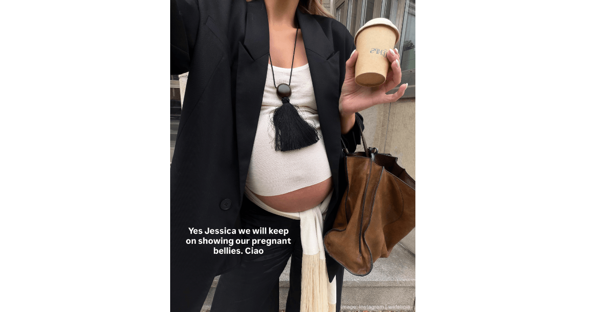 A pregnant woman showing her belly wearing a black suit jacket, black trousers, white vest and white silk fringed scarf under her bump
